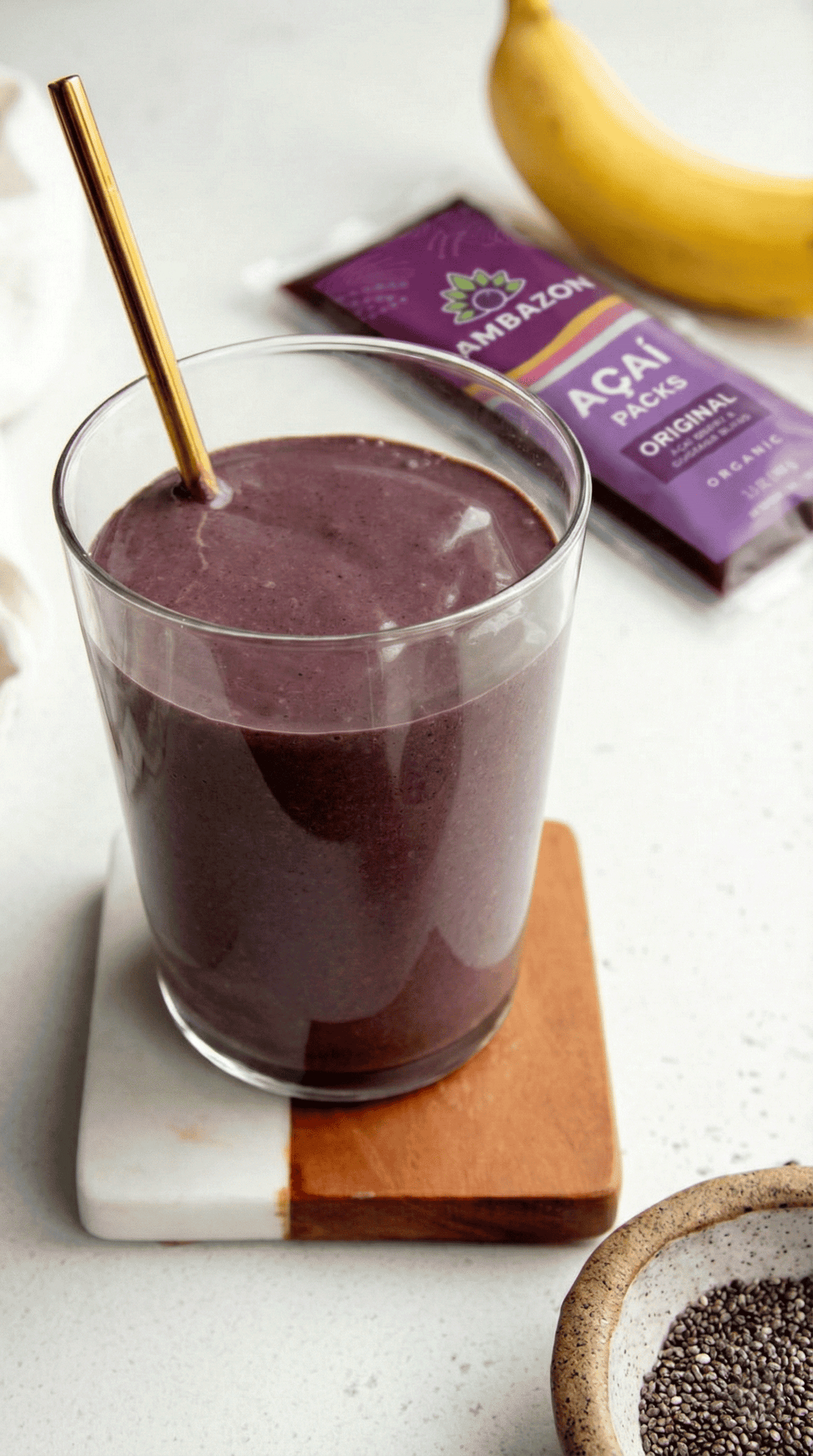 A glass of purple acai smoothie with a gold straw, sitting next to a SAMBAZON Frozen Acai Pack – Original and a banana.